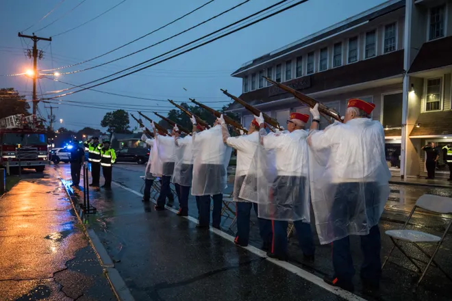 A line of Marine Corps League members lined up on a street with riffles to do a holiday tribute in the rain with their ponchos on.