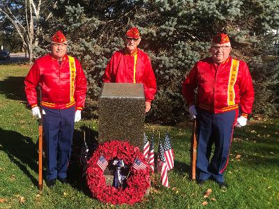 Three Marine Corps League members stand in their red silk jackets and covers (hats) and blue pants around a monument with a wreath.